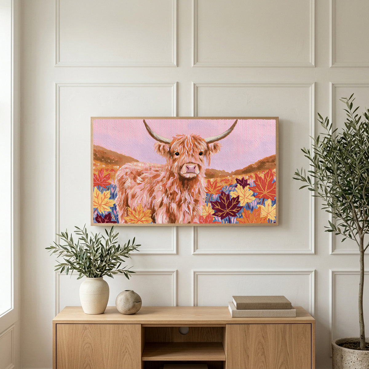 Highland cow in fall leaves art on a TV above a light oak sideboard with olive branches in a bright living room