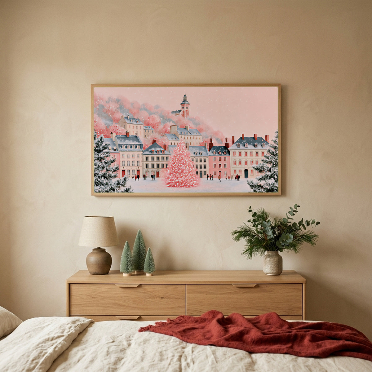 Pink Christmas village TV art above an oak dresser with small bottle-brush trees in a calm bedroom