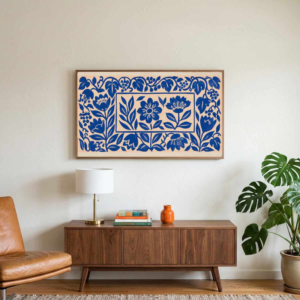 Blue paper cut flowers art on a TV above a walnut credenza with a brass lamp and a monstera plant
