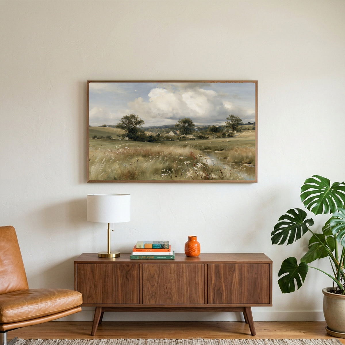 Countryside meadow and cottage art on a TV above a walnut credenza with a brass lamp and a monstera plant