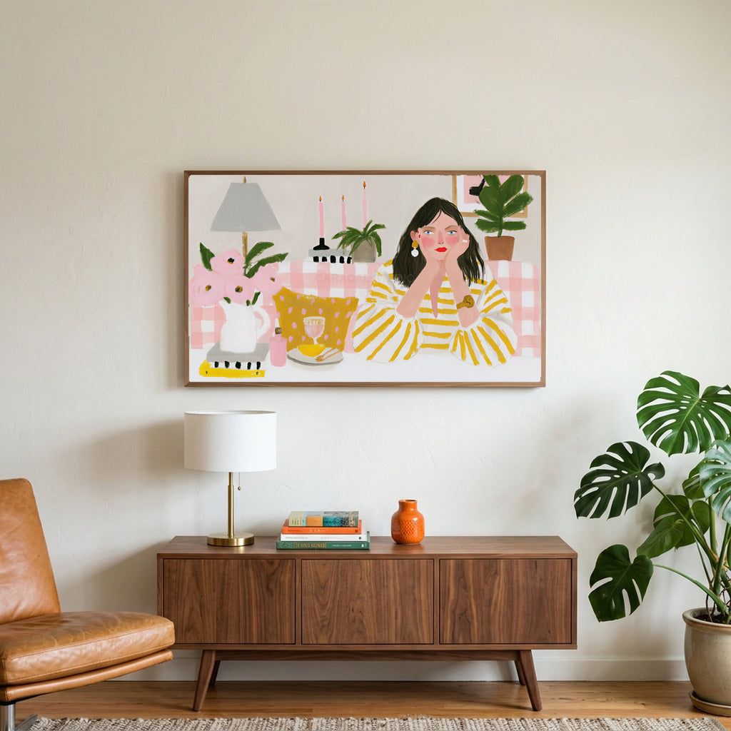 Woman at a pink table art on a TV above a walnut credenza with a brass lamp and a monstera plant